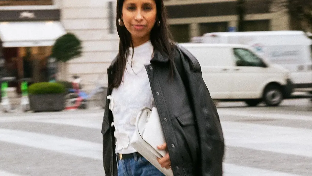 Stylische Frau kombiniert eine weiße Bluse mit Jeans und Lederjacke. Sie trägt eine weiße Tasche und goldene Ohrringe.