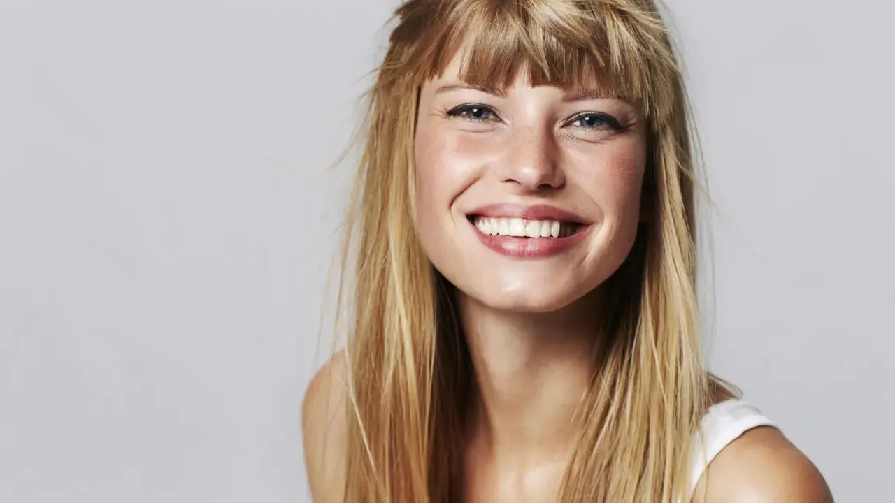 Strahlendes Lächeln einer jungen Frau mit einer lässigen Pony Frisur und langen blonden Haaren.