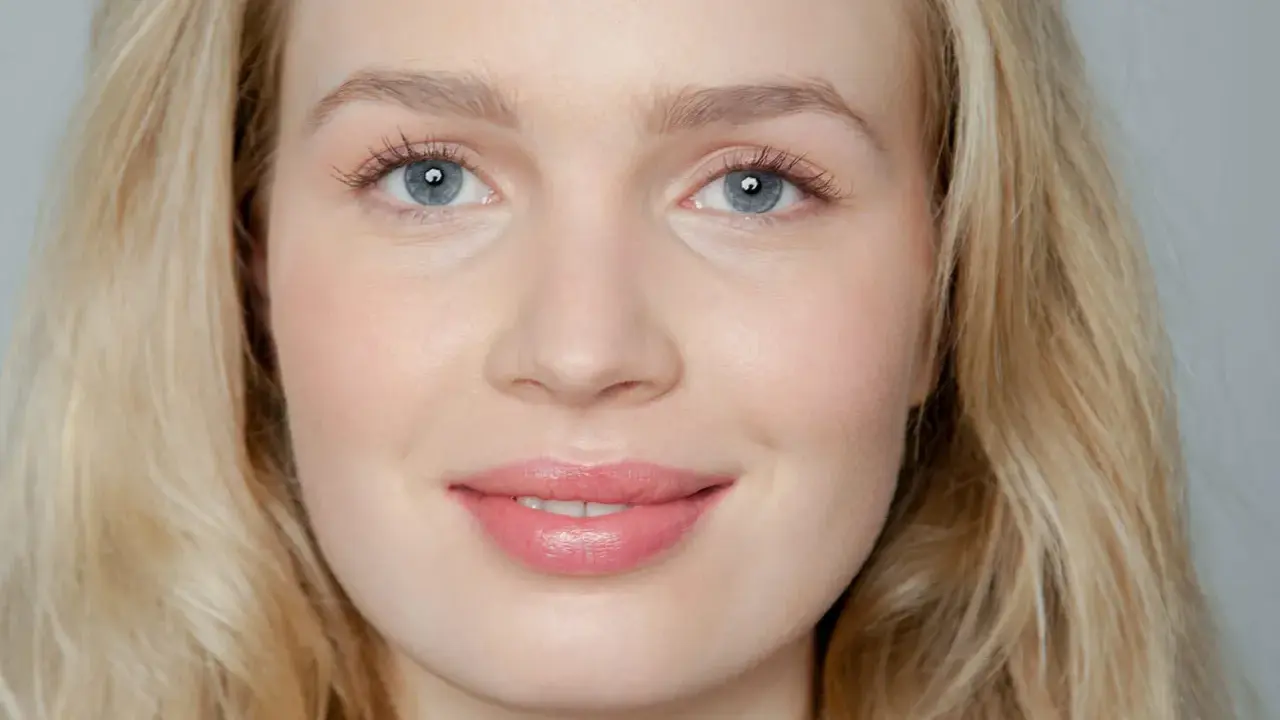 Blonde Frau mit blauen Augen und rosafarbenen Lippen, die frisch aussieht.
