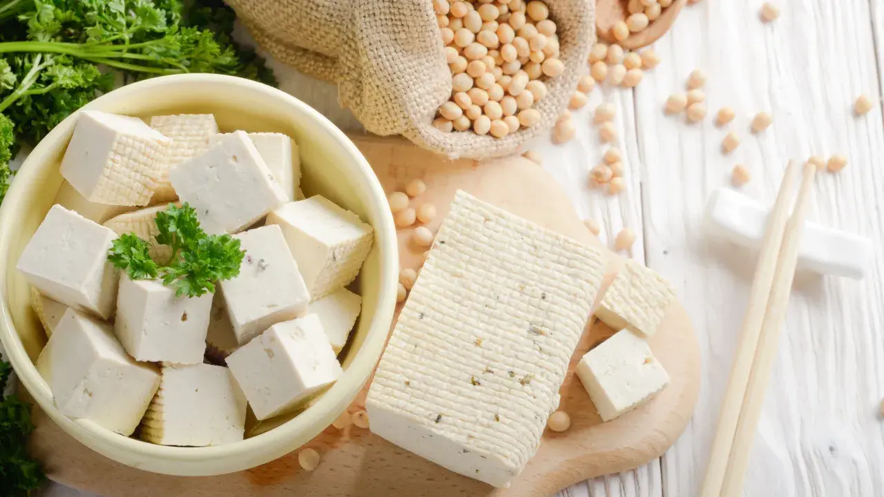 Würfel und ein Block Tofu liegen auf einem Holzbrett. Sojabohnen in einem Sack und Petersilie deuten auf die tofu inhaltsstoffe hin.