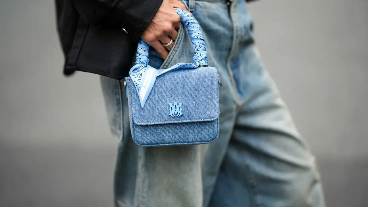 Ein hellblaues Jeans-Täschchen mit einem quadratischen Tuch binden, das den Griff ziert. Das Tuch hat ein Paisleymuster und den Schriftzug "AMIRI".