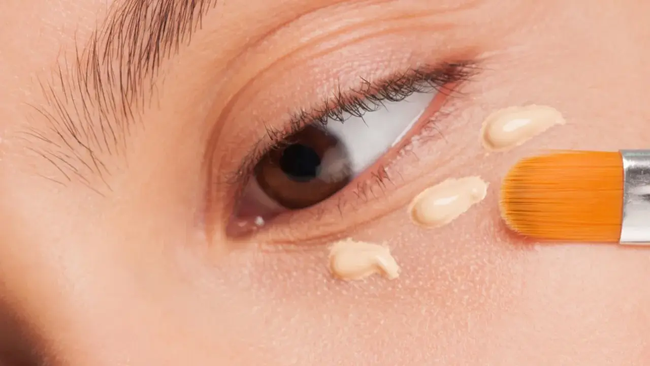 Nahaufnahme eines Auges, auf das mit einem Pinsel Concealer aufgetragen wird. Der Concealer setzt sich ab und bildet kleine Tupfen.
