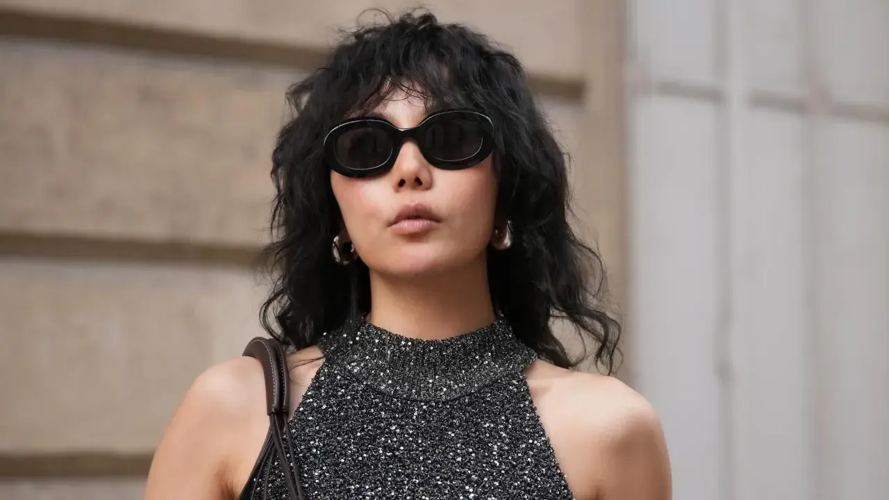 Frau mit schwarzem Wolf Cut Locken, Sonnenbrille und glitzerndem Top.
