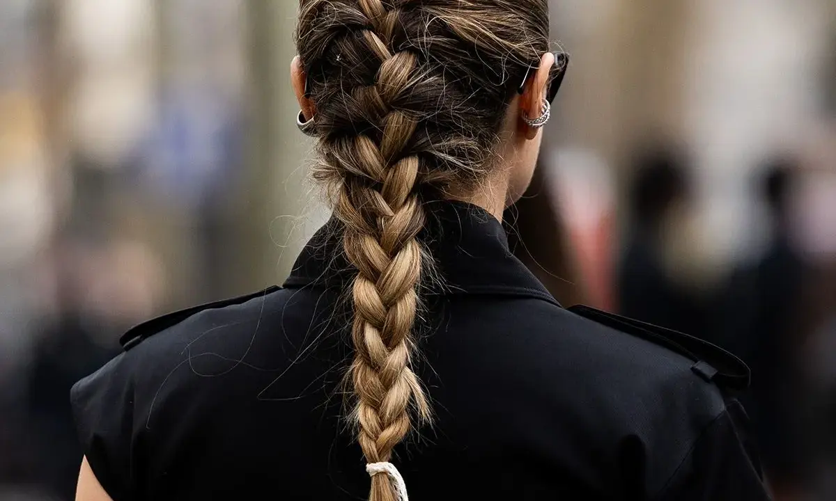 Eine Frau mit einem kunstvollen french braid, das elegant über ihre Schulter fällt. Sie trägt einen schwarzen Blazer und auffällige Ohrringe.