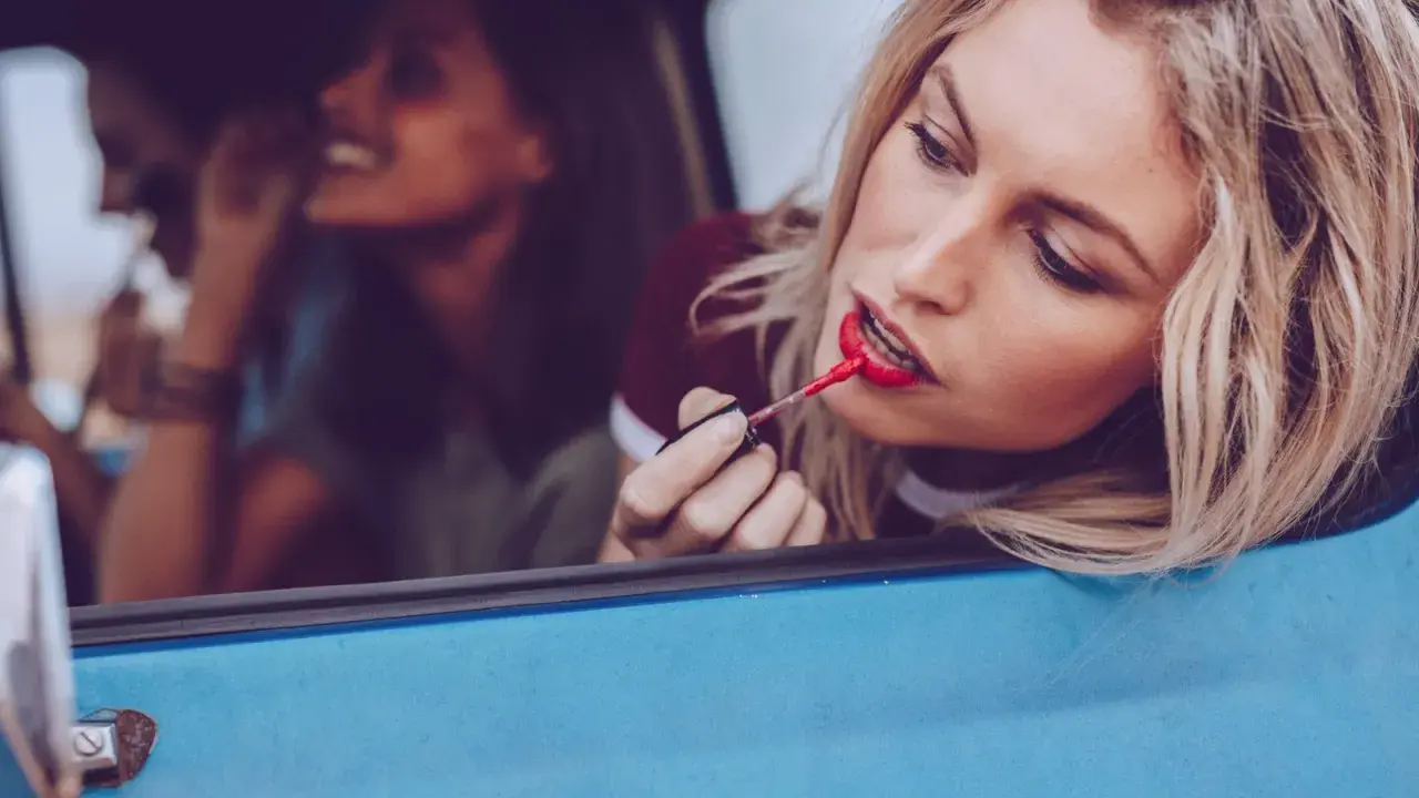 Blonde Frau mit rotem Lippenstift im Auto, um ihr Make-up aufzufrischen. Freundinnen im Hintergrund.