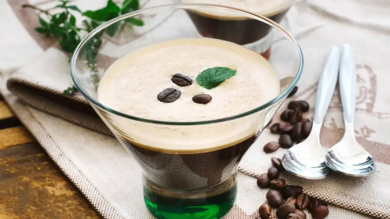 Ein Glas italienischer Eiskaffee mit Kaffeebohnen und Minze garniert, daneben Löffel und Kaffeebohnen auf einem Tuch.