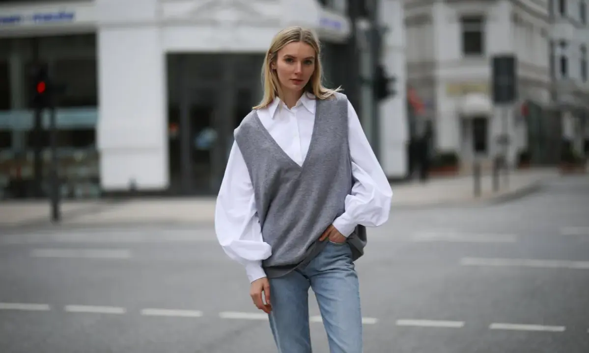 Stylische Frau in grauem Pullunder, weißer Bluse und Jeans. So lässt sich ein Pullunder kombinieren für einen lässigen Look.