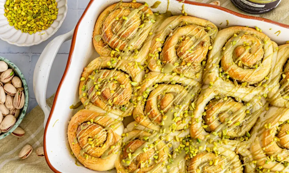 Frisch gebackene Pistazienschnecken, beträufelt mit Pistaziencreme und bestreut mit gehackten Pistazien. Daneben Schalen mit ganzen und gehackten Pistazien.