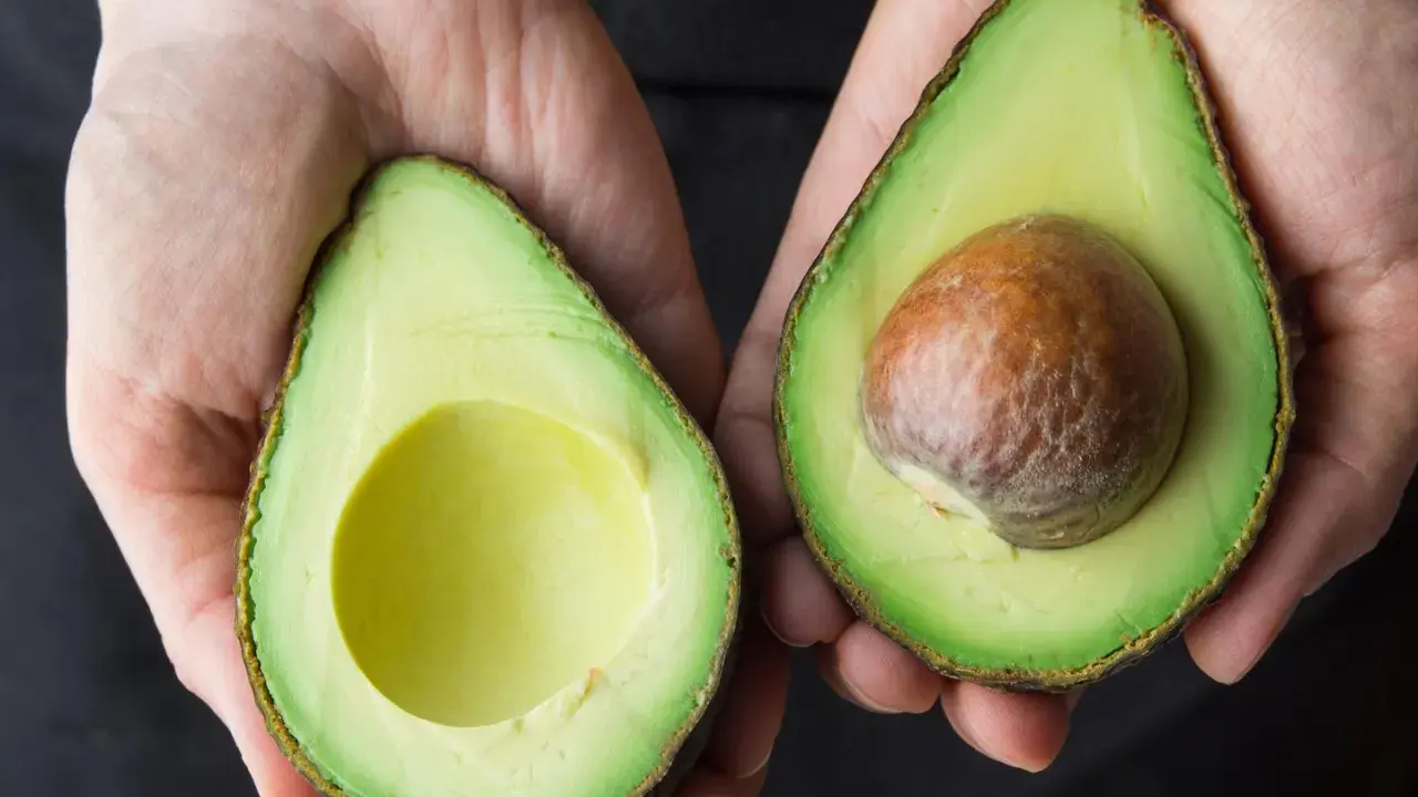 Hände halten zwei Avocadehälften. Eine Avocado mit Kern, die andere leer. Manche sagen, Avocado sei krebserregend, aber das stimmt nicht.
