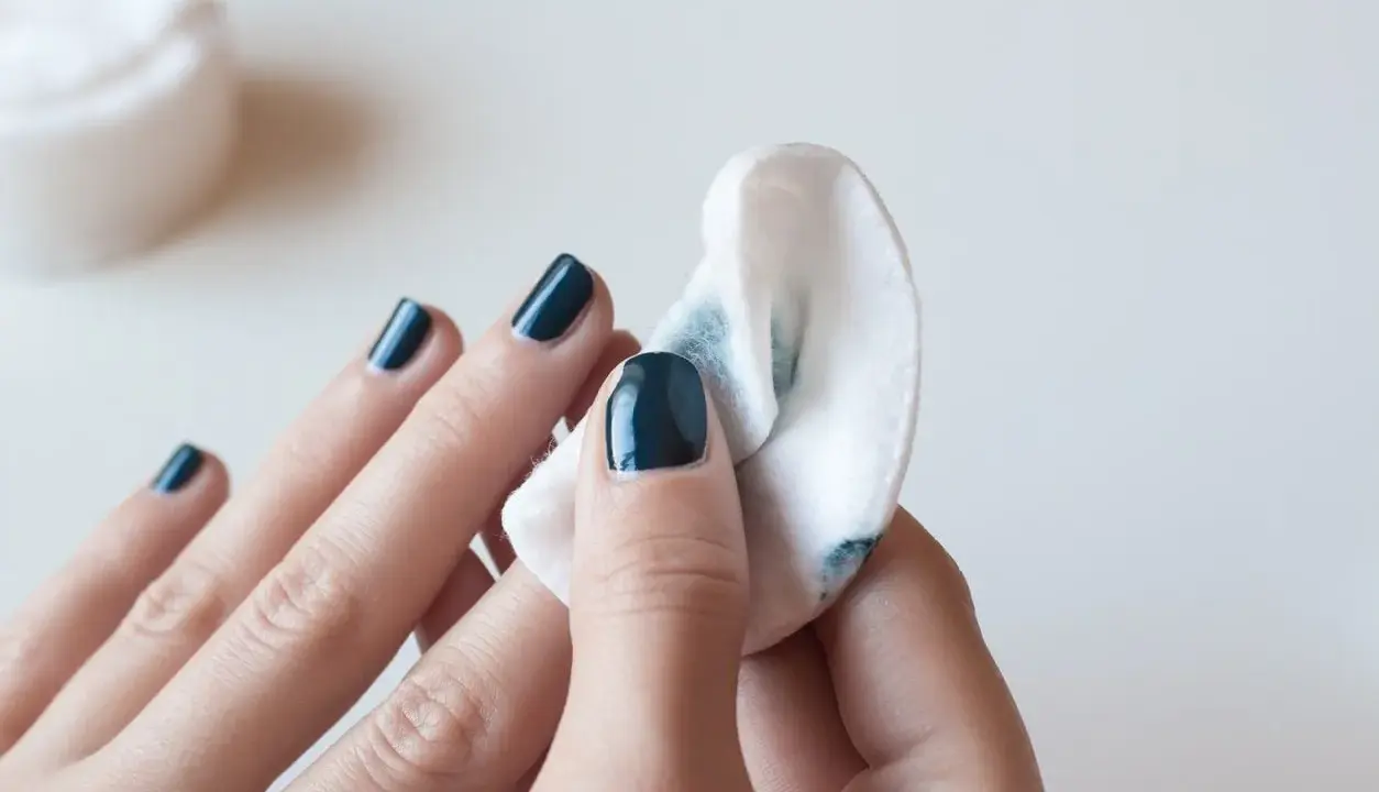 Eine Hand mit dunkelblauem Nagellack wird mit einem Wattepad abgewischt, um den Nagellack zu entfernen.