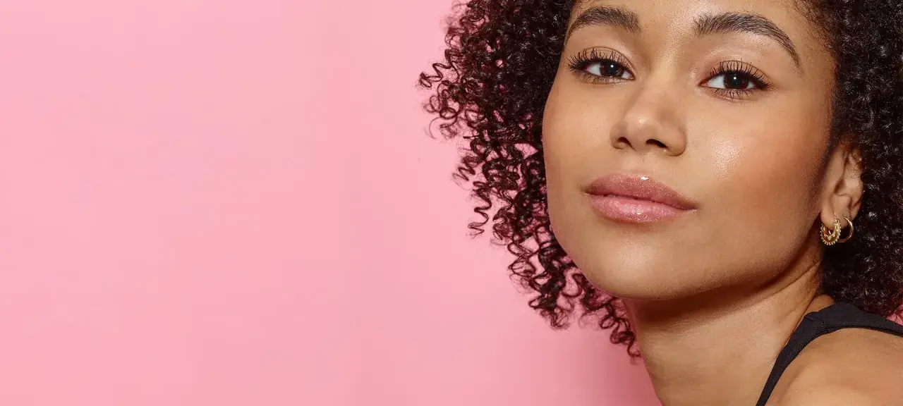 Ein Porträt einer jungen Frau mit vollen, glänzenden Lippen und lockigem Haar vor einem rosafarbenen Hintergrund. Ein Hauch von Make-up, das 50 Jahre Erfahrung widerspiegelt.