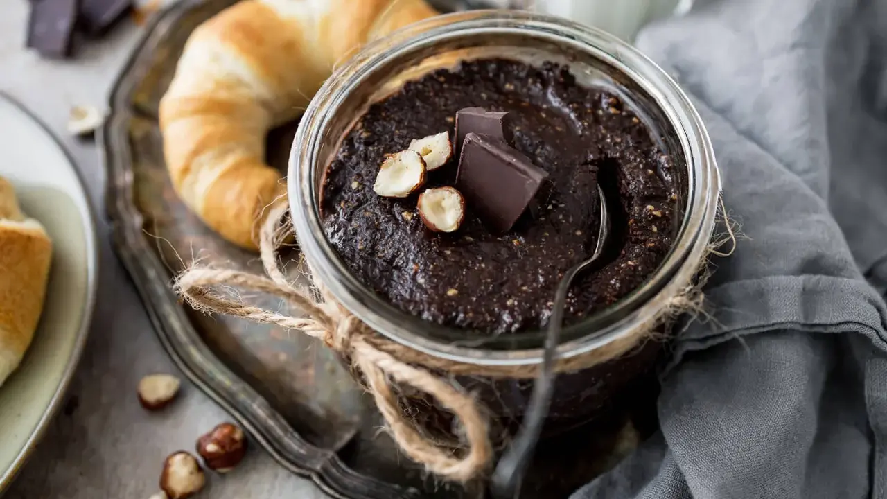 Selbstgemachte Haselnusscreme im Glas, garniert mit Schokostückchen und Nüssen, neben einem Croissant. Perfekt zum Haselnusscreme selber machen!