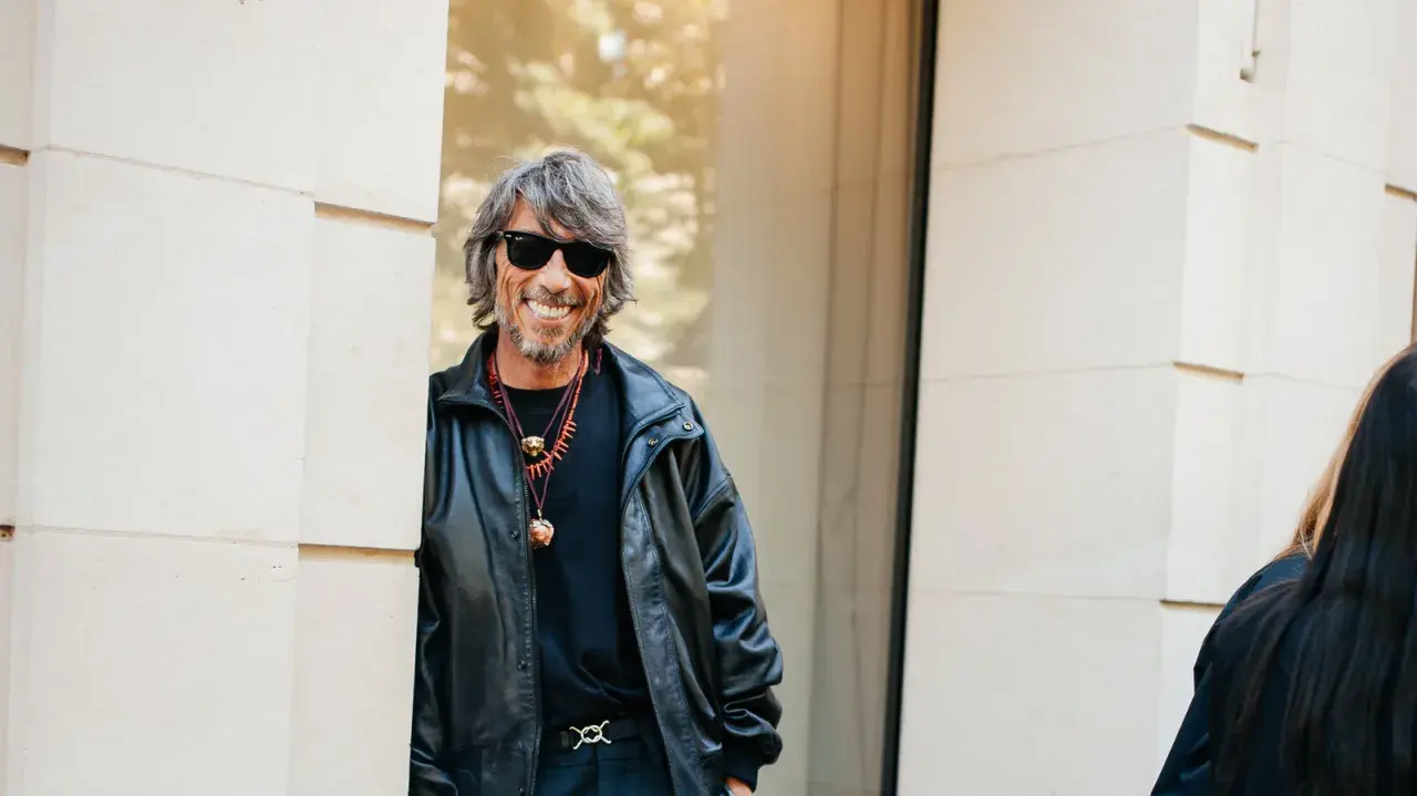 Ein Mann mit grauen Haaren und Sonnenbrille, gekleidet in eine Lederjacke, lächelt. Er könnte ein Balenciaga Designer sein, der seinen Stil zur Schau stellt.