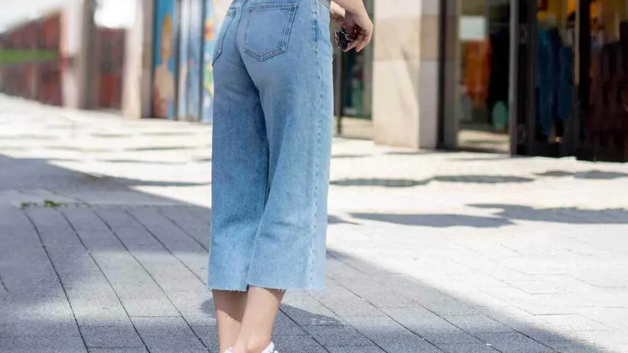 Styling culotte jeans: Ein lässiger Look mit hellblauen Jeans, weißem Shirt und Sneakern auf gepflastertem Weg.
