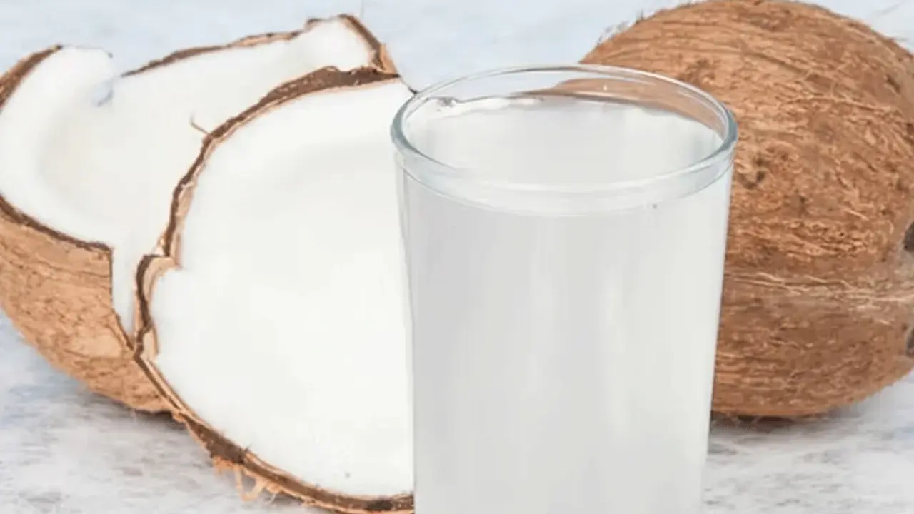Ein Glas erfrischendes Kokoswasser neben einer ganzen und einer halbierten Kokosnuss. Dieses natürliche Getränk ist bekannt dafür, dass Kokoswasser gesund ist.