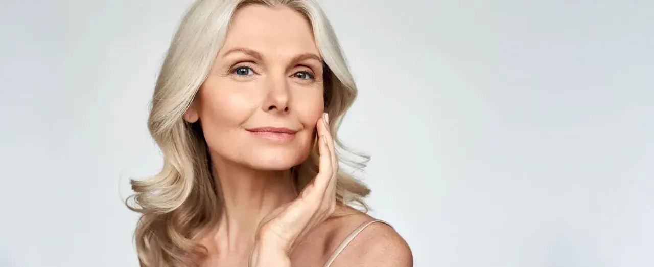 Eine Frau mit blonden Haaren berührt sanft ihr Gesicht. Ihr Make-up setzt sich in den feinen Falten ab, was ihrer Haut Tiefe verleiht.