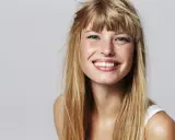 Strahlendes Lächeln einer jungen Frau mit einer lässigen Pony Frisur und langen blonden Haaren.