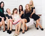 Sechs Frauen in eleganten Kleidern, die meisten davon schwarz, sitzen auf einer Bank. Eine Brautjungfer in Weiß hält ein rosa Kissen. Ein stilvolles Bild für eine Hochzeit.