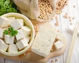 Würfel und ein Block Tofu liegen auf einem Holzbrett. Sojabohnen in einem Sack und Petersilie deuten auf die tofu inhaltsstoffe hin.