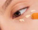 Nahaufnahme eines Auges, auf das mit einem Pinsel Concealer aufgetragen wird. Der Concealer setzt sich ab und bildet kleine Tupfen.