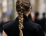 Eine Frau mit einem kunstvollen french braid, das elegant über ihre Schulter fällt. Sie trägt einen schwarzen Blazer und auffällige Ohrringe.