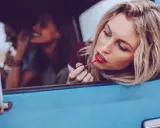 Blonde Frau mit rotem Lippenstift im Auto, um ihr Make-up aufzufrischen. Freundinnen im Hintergrund.
