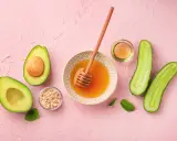 Zutaten für eine Gesichtsmaske selber machen: Avocado, Honig, Haferflocken, Gurke und Öl auf rosa Untergrund.