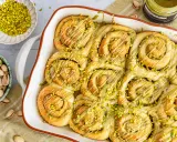 Frisch gebackene Pistazienschnecken, beträufelt mit Pistaziencreme und bestreut mit gehackten Pistazien. Daneben Schalen mit ganzen und gehackten Pistazien.