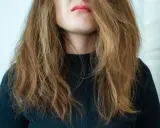 Frau mit zerzaustem, trockenem Haar, das ihr ins Gesicht fällt. Ihre Lippen sind leicht geöffnet und mit rosafarbenem Lippenstift bemalt.