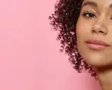 Ein Porträt einer jungen Frau mit vollen, glänzenden Lippen und lockigem Haar vor einem rosafarbenen Hintergrund. Ein Hauch von Make-up, das 50 Jahre Erfahrung widerspiegelt.