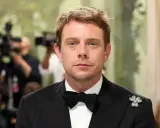 Jonathan Anderson trägt einen schwarzen Smoking mit Fliege und einem Kleeblatt-Anstecker. Im Hintergrund sind Kameras und unscharfe Personen zu sehen.