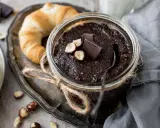 Selbstgemachte Haselnusscreme im Glas, garniert mit Schokostückchen und Nüssen, neben einem Croissant. Perfekt zum Haselnusscreme selber machen!