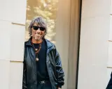 Ein Mann mit grauen Haaren und Sonnenbrille, gekleidet in eine Lederjacke, lächelt. Er könnte ein Balenciaga Designer sein, der seinen Stil zur Schau stellt.