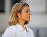 Eine Frau mit einem schicken Bob und auffälligen Dior-Sonnenbrillen. Sie trägt eine weiße Jeansjacke und Perlenschmuck.