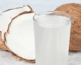 Ein Glas erfrischendes Kokoswasser neben einer ganzen und einer halbierten Kokosnuss. Dieses natürliche Getränk ist bekannt dafür, dass Kokoswasser gesund ist.