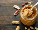 Ein Löffel cremiger Erdnussbutter ragt aus einem Glas. Umgeben von Erdnüssen und Schalen, zeigt das Bild, wie erdnussbutter gesund sein kann.