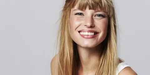 Strahlendes Lächeln einer jungen Frau mit einer lässigen Pony Frisur und langen blonden Haaren.