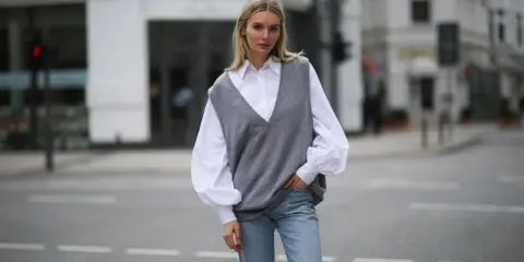 Stylische Frau in grauem Pullunder, weißer Bluse und Jeans. So lässt sich ein Pullunder kombinieren für einen lässigen Look.