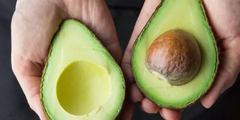 Hände halten zwei Avocadehälften. Eine Avocado mit Kern, die andere leer. Manche sagen, Avocado sei krebserregend, aber das stimmt nicht.