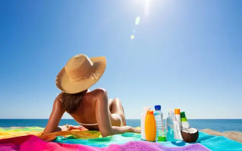 Frau mit Hut liegt am Strand, umgeben von Sonnencremes und Kokosnuss. So wird man schnell braun.