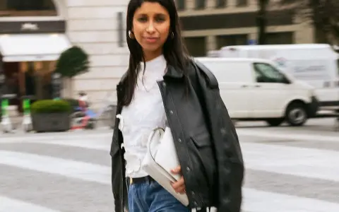 Stylische Frau kombiniert eine weiße Bluse mit Jeans und Lederjacke. Sie trägt eine weiße Tasche und goldene Ohrringe.