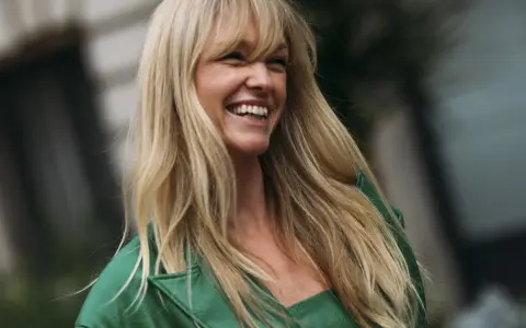 Lachende Frau mit blonden, langen Haaren und einem leichten Pony, trägt ein grünes Outfit.