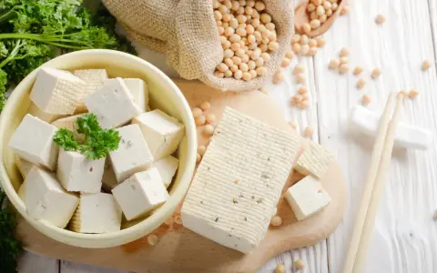 Würfel und ein Block Tofu liegen auf einem Holzbrett. Sojabohnen in einem Sack und Petersilie deuten auf die tofu inhaltsstoffe hin.