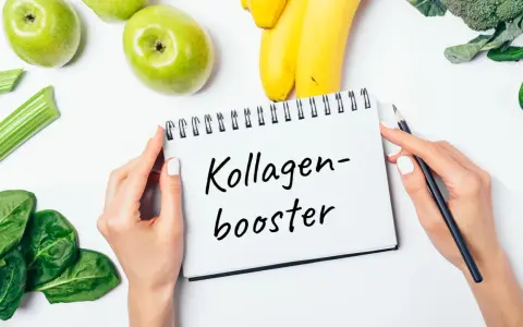 Hände halten Notizbuch mit "Kollagen-Booster". Frisches Obst und Gemüse wie Äpfel, Bananen, Sellerie, Spinat und Brokkoli liegen daneben. Eine Kollagen Lebensmittel Tabelle könnte hier inspirieren.
