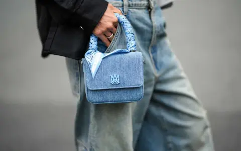 Ein hellblaues Jeans-Täschchen mit einem quadratischen Tuch binden, das den Griff ziert. Das Tuch hat ein Paisleymuster und den Schriftzug "AMIRI".