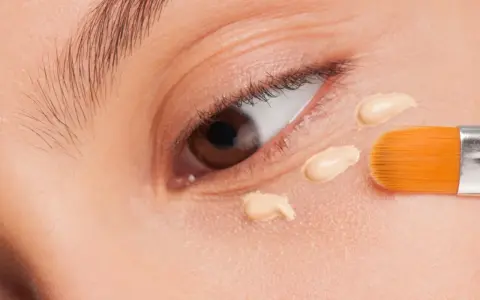 Nahaufnahme eines Auges, auf das mit einem Pinsel Concealer aufgetragen wird. Der Concealer setzt sich ab und bildet kleine Tupfen.