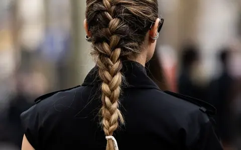 Eine Frau mit einem kunstvollen french braid, das elegant über ihre Schulter fällt. Sie trägt einen schwarzen Blazer und auffällige Ohrringe.