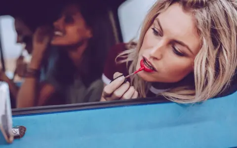 Blonde Frau mit rotem Lippenstift im Auto, um ihr Make-up aufzufrischen. Freundinnen im Hintergrund.