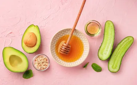 Zutaten für eine Gesichtsmaske selber machen: Avocado, Honig, Haferflocken, Gurke und Öl auf rosa Untergrund.