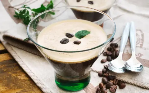 Ein Glas italienischer Eiskaffee mit Kaffeebohnen und Minze garniert, daneben Löffel und Kaffeebohnen auf einem Tuch.